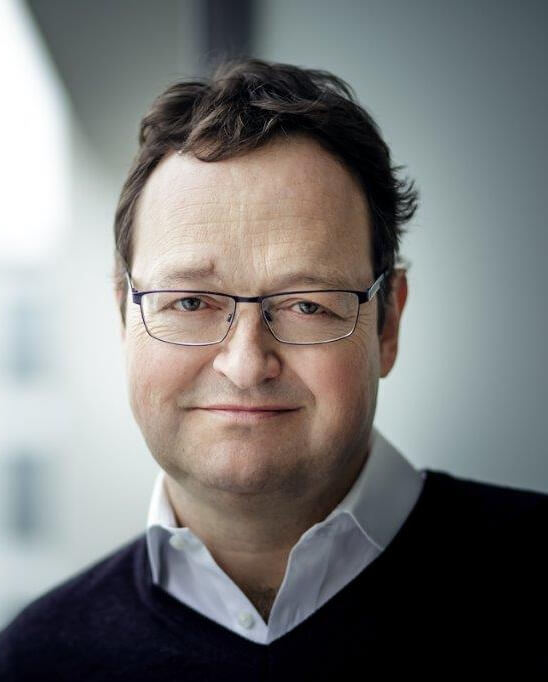 Ein lächelnder Mann mit Brille, der in einem modernen Büro steht und freundlich in die Kamera schaut.