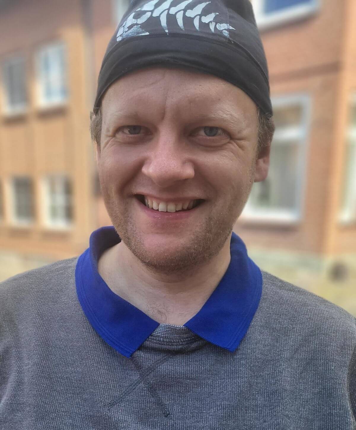 Lächelnder Mann mit schwarzer Mütze und grauem Pullover, steht vor einem Gebäude mit Fenstern.