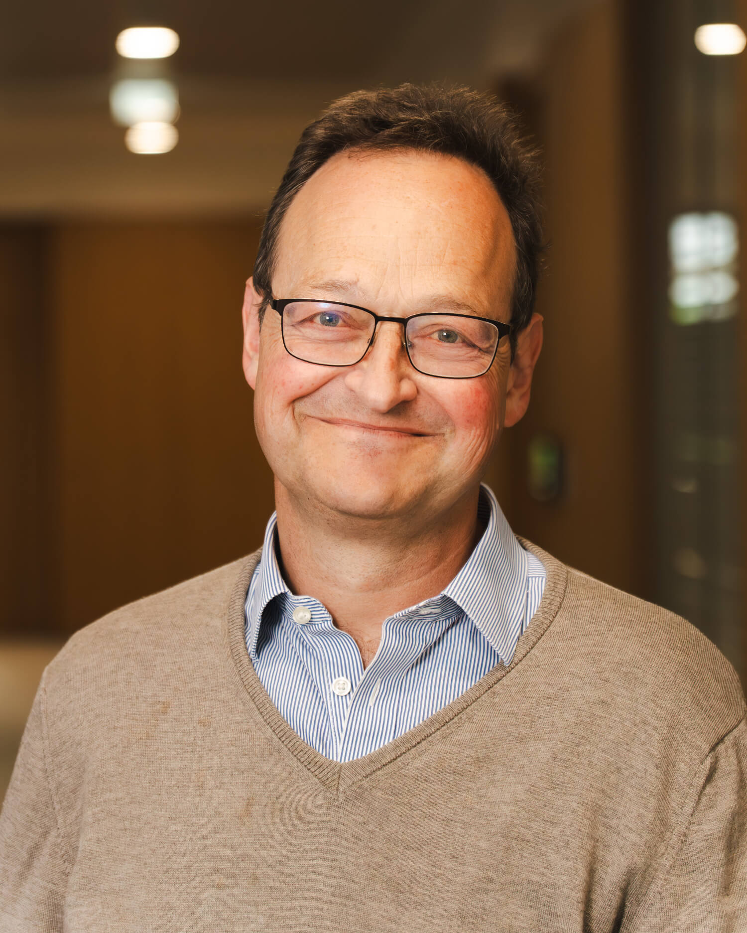 Mann mit Brille und lächelndem Gesicht, trägt einen beigen Pullover und ein gestreiftes Hemd.